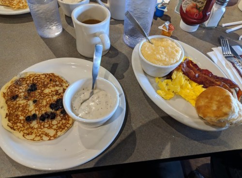 A breakfast spread featuring pancakes with blueberries, scrambled eggs, bacon, a biscuit, and a side of gravy.