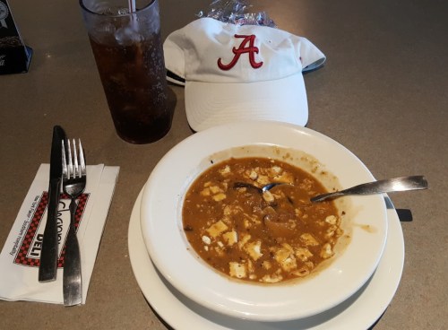 A bowl of soup with tofu, a spoon, a soft drink, and a white cap with an "A" logo on a table.