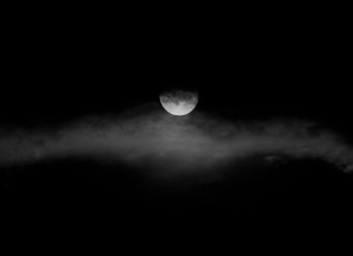A black and white image of a partially obscured moon surrounded by dark clouds against a black sky.