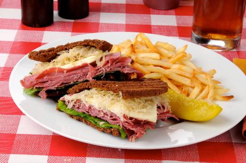 A plate with two stacked sandwiches filled with meat and sauerkraut, served with fries and a lemon wedge on a checkered tablecloth.