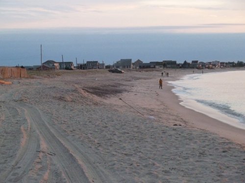 A sandy beach at dusk with a few people walking, houses in the background, and gentle waves lapping at the shore.