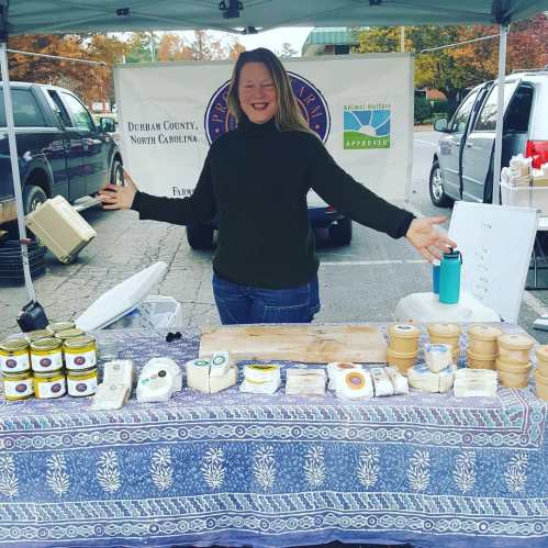 A smiling vendor stands behind a table filled with various cheeses and jars at a market under a tent.