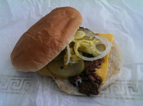 A sandwich with a beef patty, cheddar cheese, pickles, onions, and mustard on a soft bun, resting on white paper.