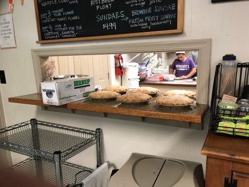 A view through a serving window showing freshly baked pies on a shelf, with a kitchen visible in the background.