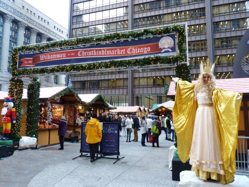 A festive market scene with a golden-draped figure welcoming visitors, surrounded by holiday stalls and decorations.