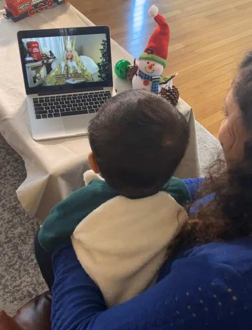A child sits on a woman's lap, watching a festive video on a laptop, with holiday decorations in the background.