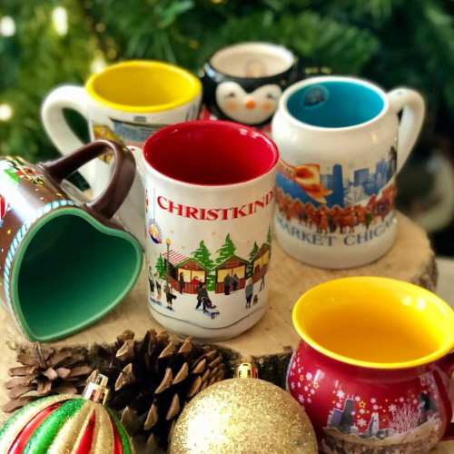 A collection of colorful holiday mugs on a wooden surface, surrounded by pinecones and ornaments.
