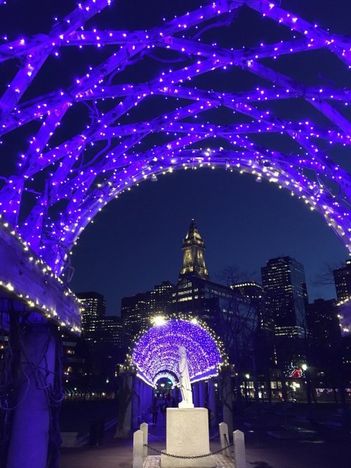 A lit archway with purple lights frames a statue, with city buildings visible in the background at dusk.