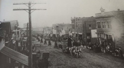A historic street scene with horse-drawn carriages, pedestrians, and early 20th-century buildings.