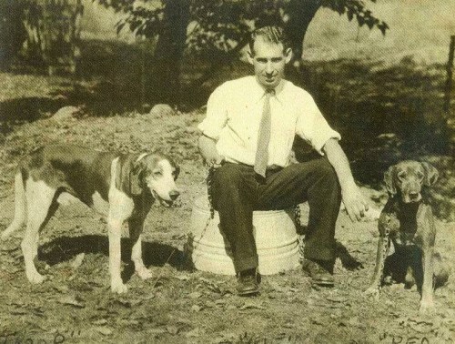 A man in a white shirt and tie sits on a bucket with two dogs beside him in a natural outdoor setting.