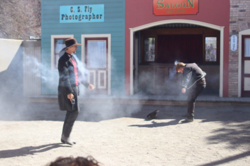 Two cowboys in a dusty street, one aiming a gun while the other bends down, with smoke swirling around them.