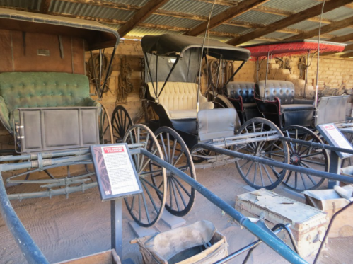 A display of vintage horse-drawn carriages in a rustic barn setting, with informational plaques nearby.