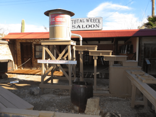 A rustic saloon with a water tower, wooden structures, and a sign reading "Total Wreck Saloon" in a desert setting.