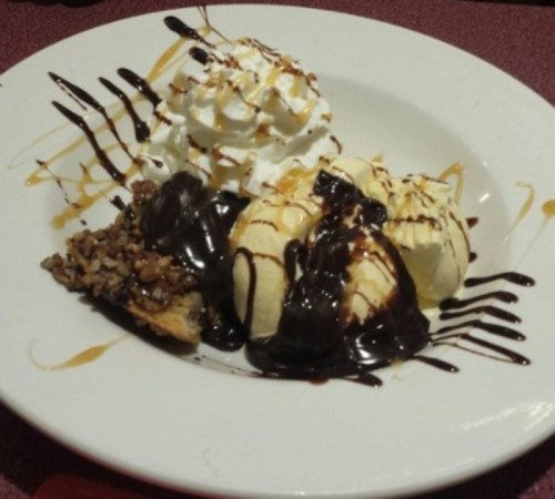 A dessert plate with three scoops of ice cream, chocolate sauce, whipped cream, and a slice of nutty pie.