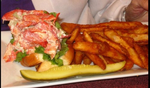 A lobster roll with lettuce on a bun, served with crispy fries and a pickle on a white plate.