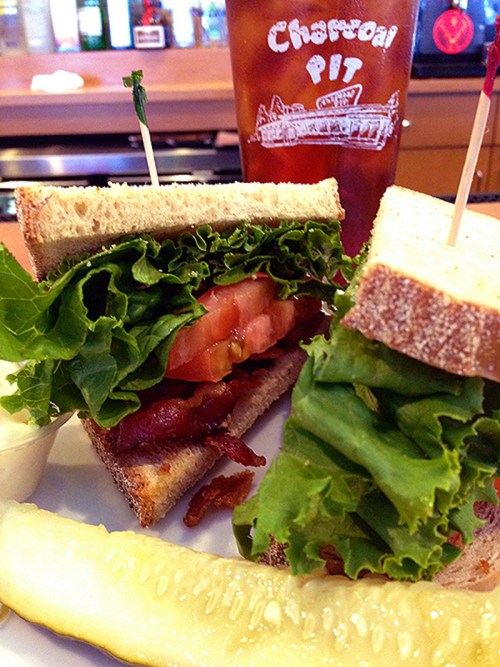 A close-up of a sandwich with lettuce, tomato, and bacon, served with a pickle and iced tea in the background.