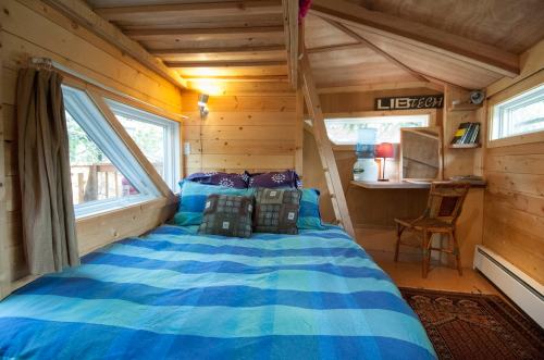 Cozy wooden interior of a tiny house with a bed, desk, and large windows, featuring blue and green bedding.