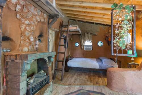 Cozy rustic interior featuring a wooden loft bed, stone fireplace, and unique circular windows. Warm earthy tones.