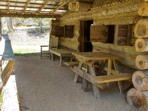 A rustic log cabin with a wooden table and chairs, surrounded by nature and a gravel floor.