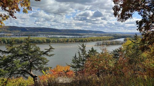 A scenic view of a river winding through lush greenery and autumn-colored trees under a cloudy sky.