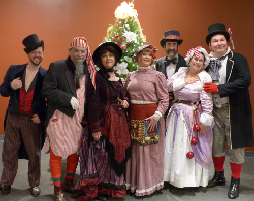 A group of eight people in festive Victorian costumes poses in front of a decorated Christmas tree.