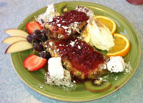 A colorful plate of French toast topped with raspberry sauce, served with fruit, whipped cream, and powdered sugar.