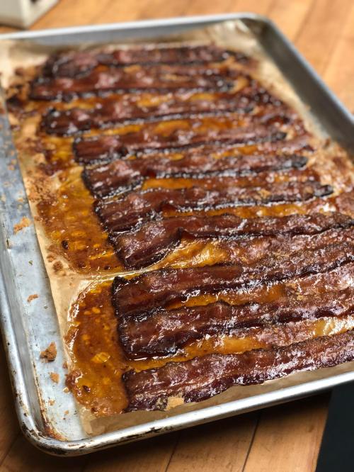 A baking sheet with crispy, caramelized bacon strips arranged neatly, with some glaze pooling around them.