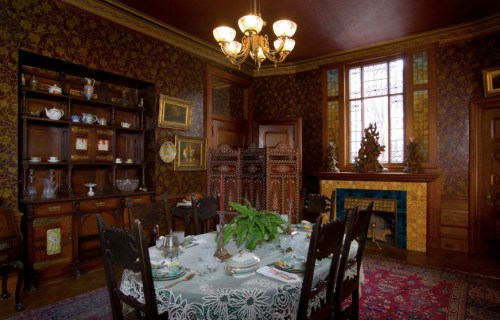 A vintage dining room with a wooden table, ornate chairs, a patterned wallpaper, and a decorative fireplace.