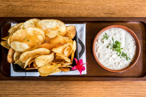 A wooden tray with a serving of crispy potato chips and a bowl of creamy dip, garnished with herbs and a flower.