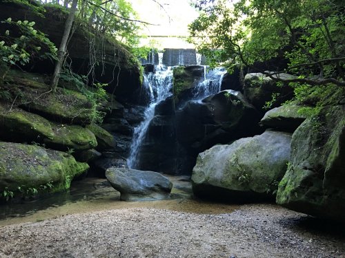 A serene waterfall cascades over moss-covered rocks, surrounded by lush greenery and a sandy riverbank.