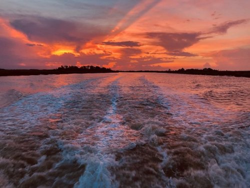 A vibrant sunset over water, with colorful clouds and rippling waves trailing behind a boat.