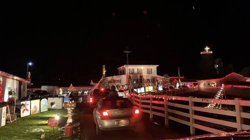 Cars lined up at a festive display of Christmas lights and decorations at night, with a large cross illuminated in the background.