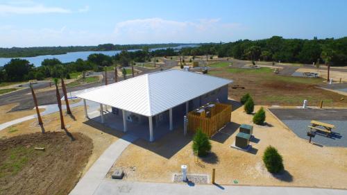 A modern building with a metal roof surrounded by open land and trees, near a body of water under a clear blue sky.