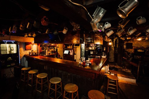 A cozy bar interior with wooden stools, a long counter, and hanging mugs, illuminated by warm lighting.