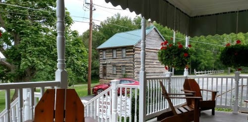 A view from a porch with wooden chairs, featuring a log cabin and lush greenery in the background.