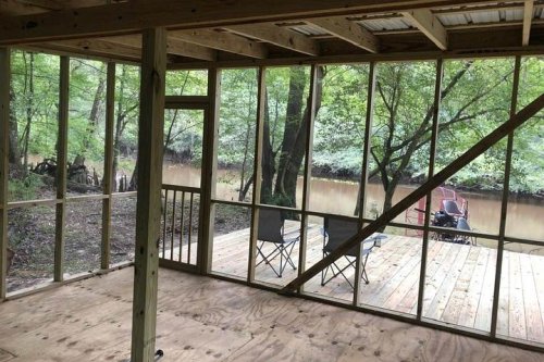 Interior view of a screened porch overlooking a river, with wooden flooring and two chairs facing the water.