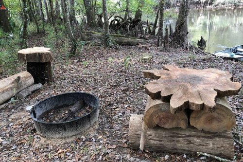 A rustic campsite by a river, featuring a fire pit, wooden seating, and a natural forest setting.