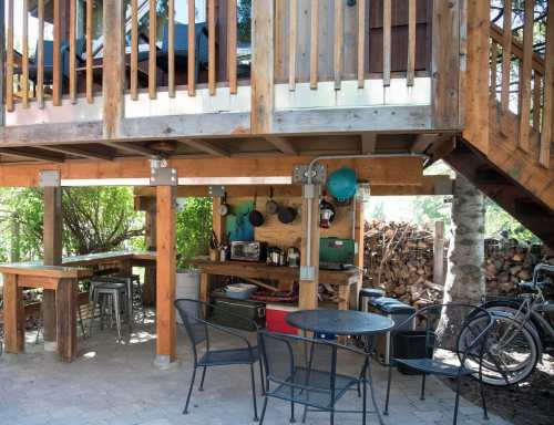 A cozy outdoor kitchen area with wooden beams, a table, chairs, and stacked firewood nearby.