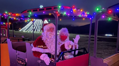 Santa and Mrs. Claus wave from a decorated train, surrounded by colorful holiday lights in a festive nighttime setting.
