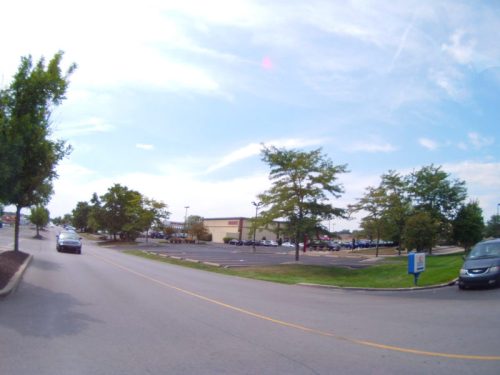A wide street with trees on the sides, leading to a parking lot and a building in the background under a blue sky.