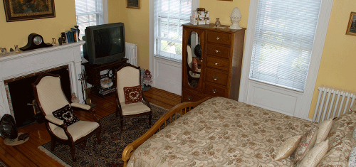 Cozy bedroom with a bed, two chairs, a dresser, a TV, and large windows, featuring warm yellow walls and wooden floors.