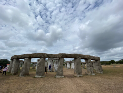 Visitors explore a stone circle structure under a cloudy sky, with grassy ground and distant trees.