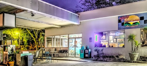 A colorful burger station at night, featuring neon lights and outdoor seating surrounded by trees.