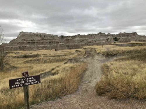 A dirt trail leads through grassy terrain with rocky formations in the background, warning sign for rough terrain visible.