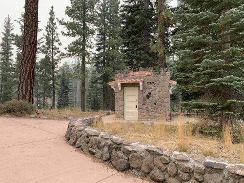 A small stone building with a white door, surrounded by tall trees and a gravel path in a misty forest setting.