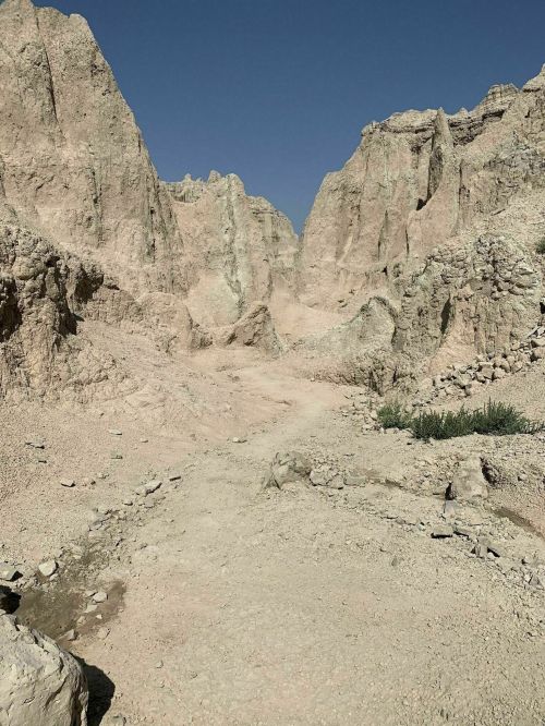 A dry, rocky landscape with steep, eroded cliffs under a clear blue sky. Sparse vegetation is visible in the foreground.