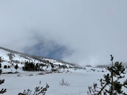 A snowy landscape with low clouds and a hint of blue sky, surrounded by trees and distant mountains.