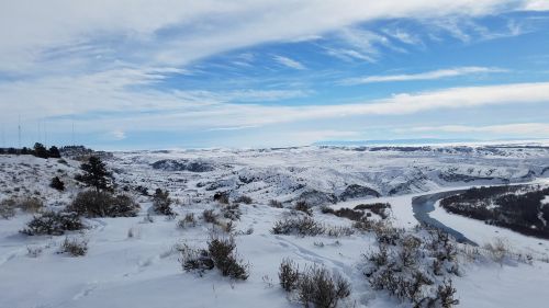 Snow-covered landscape with rolling hills, a winding river, and a blue sky with scattered clouds.