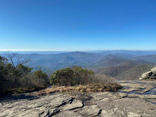 A panoramic view of rolling mountains under a clear blue sky, with rocky foreground and sparse vegetation.