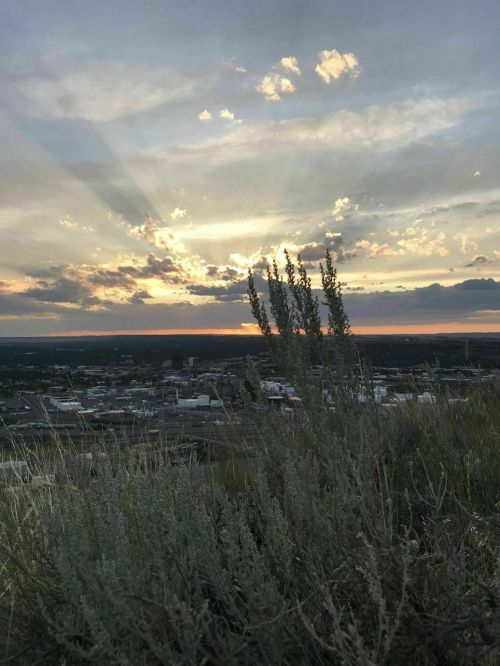A sunset over a cityscape, with rays of light breaking through clouds and sagebrush in the foreground.